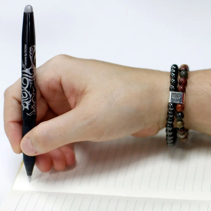 A black pen with a silver swirl design held above a lined notebook next to a magnetic gemstone bracelet with picasso jasper