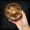 A hand holds a round mango wood incense burner disc with seven carved chakra symbols and a brass incense hole