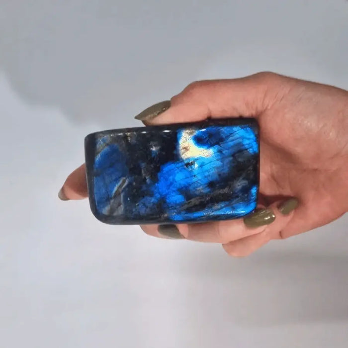 A hand holds a polished blue madagascar labradorite stone 8-10cm with swirling patterns and an iridescent sheen