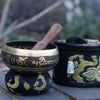 A dark bronze mini singing bowl with gold sanskrit inscriptions and wooden mallet on a black cushion with gold patterns