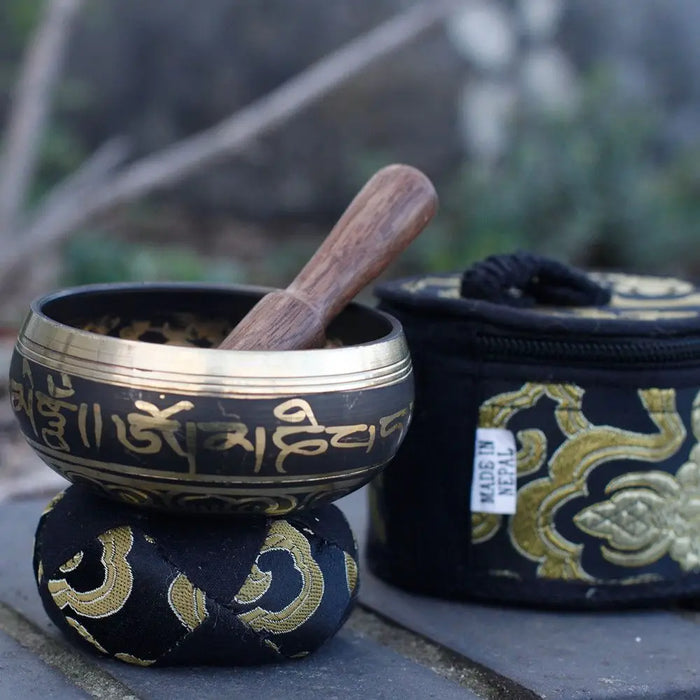 A dark bronze mini singing bowl with gold sanskrit inscriptions and wooden mallet on a black cushion with gold patterns