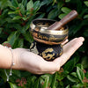 Black mini singing bowl with gold trim and engraved sanskrit script with wooden mallet resting inside