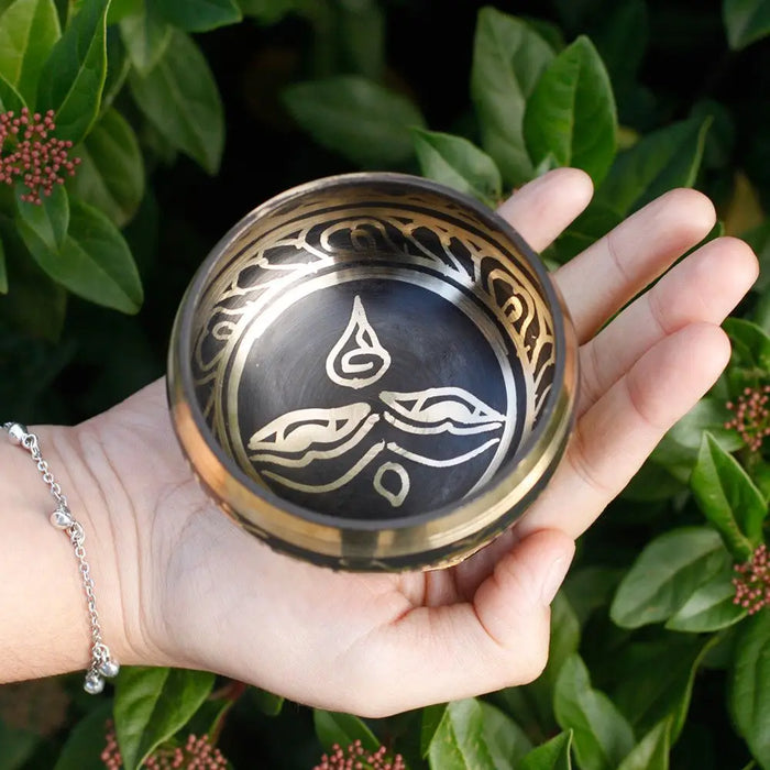 A hand holds a small dark bronze mini singing bowl with gold engravings and lotus petal design