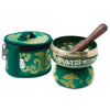 Green mini singing bowl with gold trim and sanskrit script resting on an ornate cushion with wooden mallet