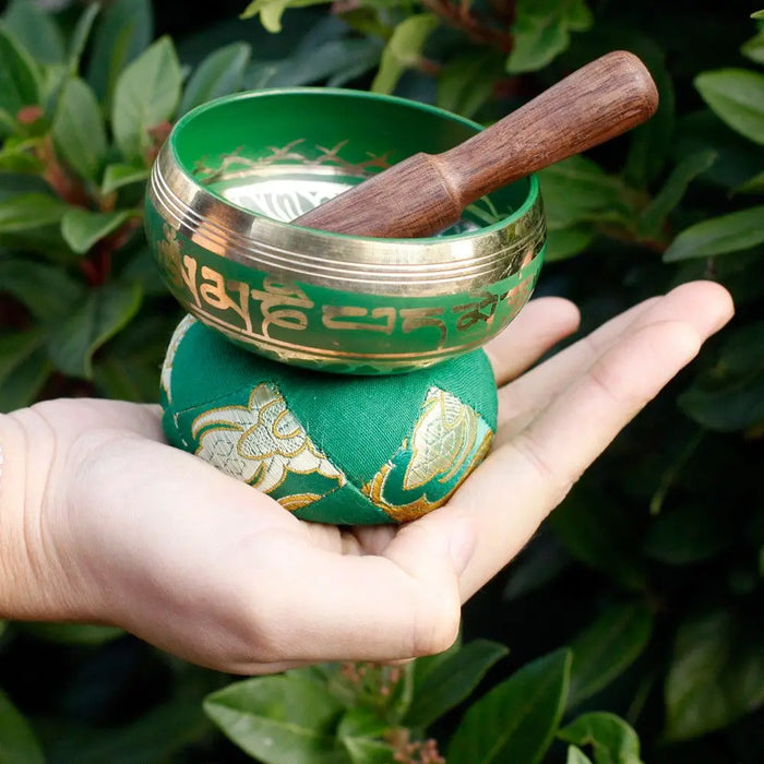 Green mini singing bowl with gold trim and engraved sanskrit resting on a matching green embroidered cushion