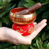 A red and gold mini singing bowl with engraved Sanskrit script resting on a matching embroidered cushion