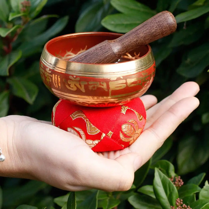 A red and gold mini singing bowl with engraved Sanskrit script resting on a matching embroidered cushion