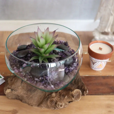 A clear glass open bowl with a green succulent and purple tips set on dark river stones and crushed amethyst on gamal root