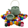 A sage green ceramic teapot with gold handle and lid knob sits on a colorful multi-colour felt ball centre mat