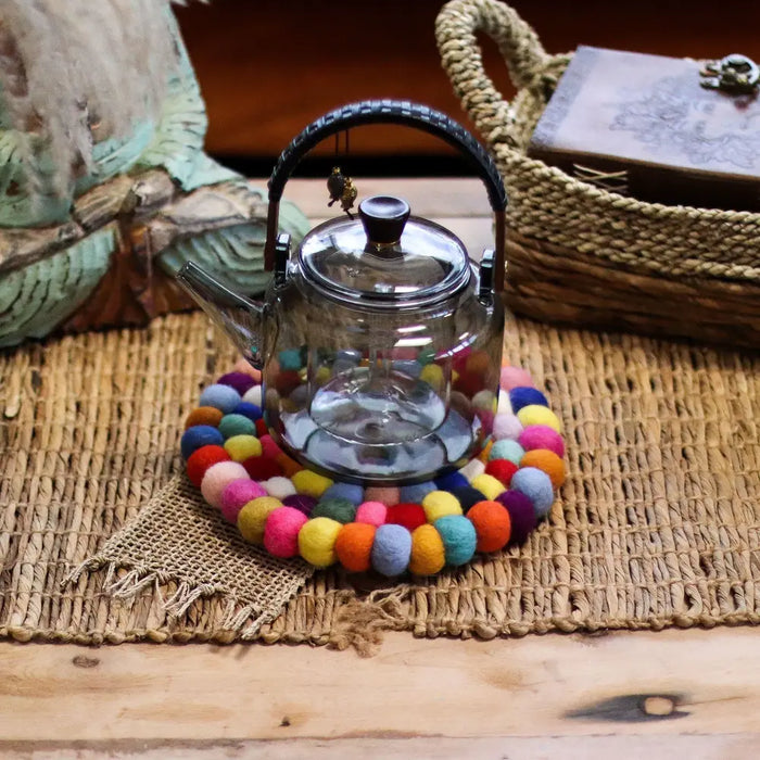 Clear glass teapot with black woven handle on a round multi-colour felt ball placemat trivet