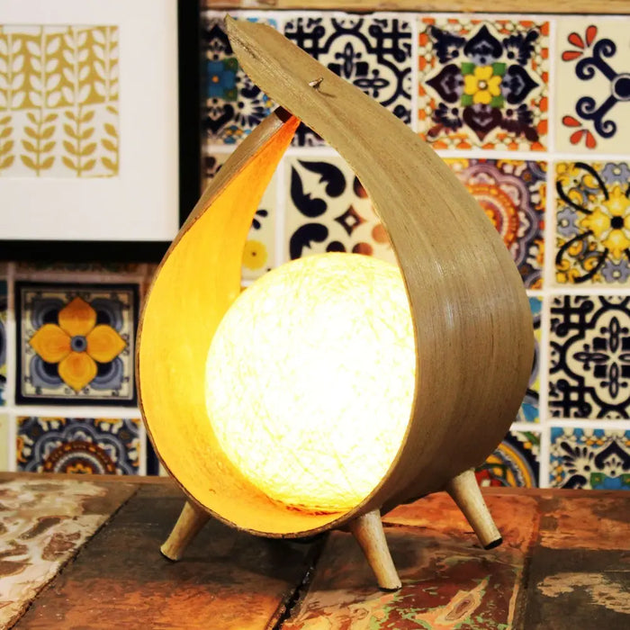 A warm glowing spherical coconut lamp with a light brown wooden shell and three slender legs natural loop design