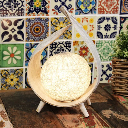 A warm glowing coconut lamp with a textured surface in a whitewashed wooden frame with three slender legs