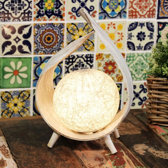 A warm glowing coconut lamp with a textured surface in a whitewashed wooden frame with three slender legs
