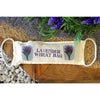 A beige linen lavender wheat bag with purple floral embroidery and white rope handles filled with golden grains