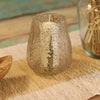 A clear glass candle holder with speckled texture and embossed fern design holding a white wax candle made of recycled glass
