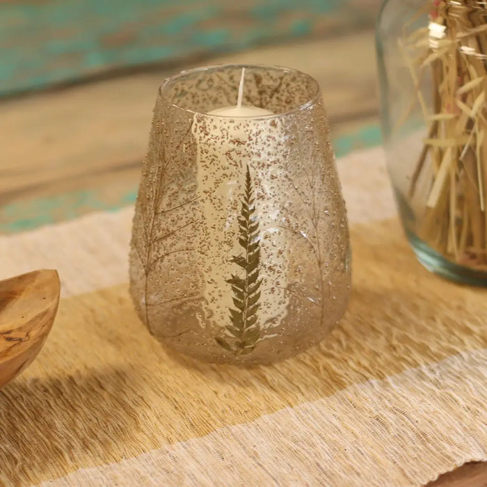 A clear glass candle holder with speckled texture and embossed fern design holding a white wax candle made of recycled glass