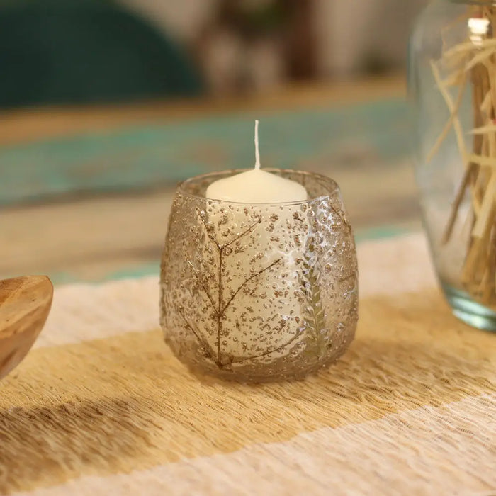 A clear recycled glass candle holder with dried floral designs holding a white pillar candle natural floral pino style