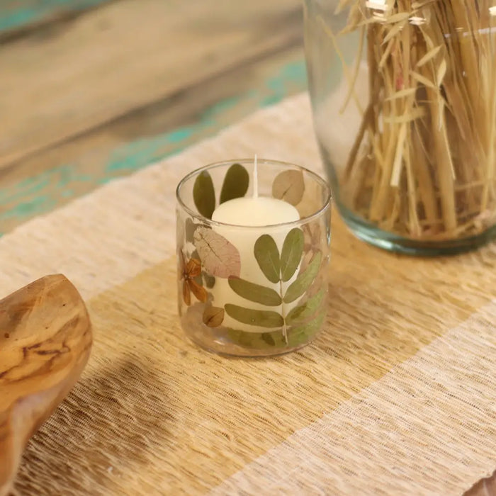 Clear glass candle holder with pressed green and brown leaves made from recycled glass holding a white pillar candle