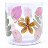 A clear glass candle holder with preserved pink and green leaves and a golden flower made from recycled glass