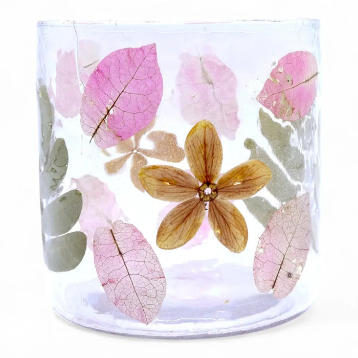 A clear glass candle holder with preserved pink and green leaves and a golden flower made from recycled glass