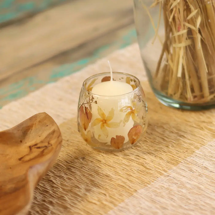 A clear recycled glass candle holder with embedded dried yellow and brown flowers holding a white wax candle