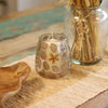 A clear glass candle holder with embedded golden and brown leaves holding a white wax candle made from recycled glass