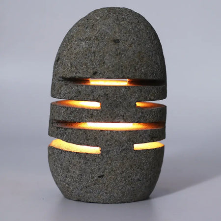 Dark gray riverstone candle holder with horizontal cutouts emitting warm orange light natural volcanic stone