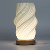 A modern new wave lamp with a white sculpted twisted body and natural wood base softly glowing on a table