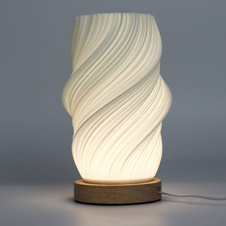A modern new wave lamp with a white sculpted twisted body and natural wood base softly glowing on a table