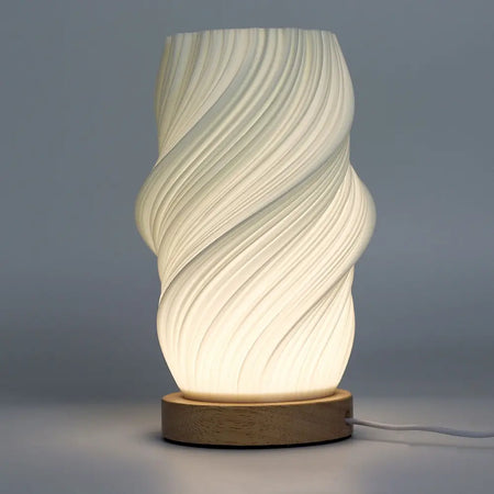 A modern new wave lamp with a white sculpted twisted body and natural wood base softly glowing on a table