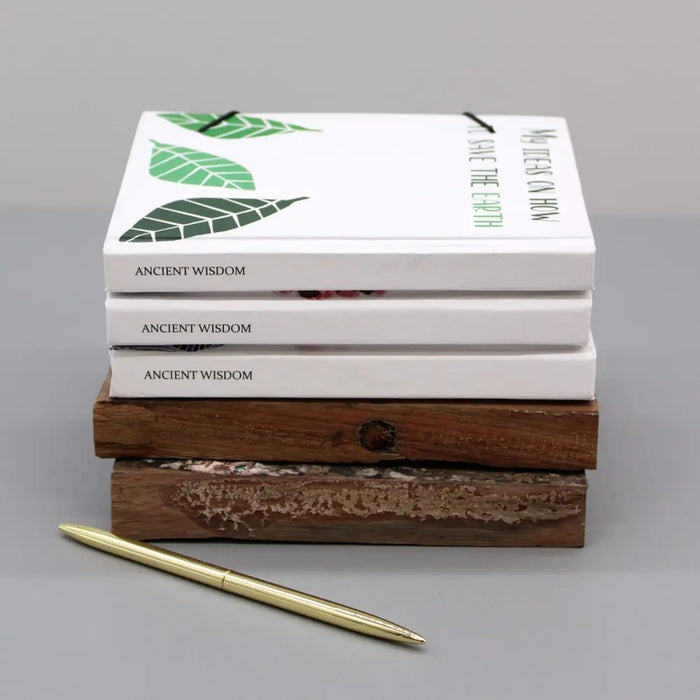 Stack of three white hardcover notebooks with green leaf designs and strap resting on rustic wooden blocks
