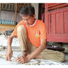 A man in an orange marimas t-shirt kneels working with thick white rope on a natural black and white seagrass rug