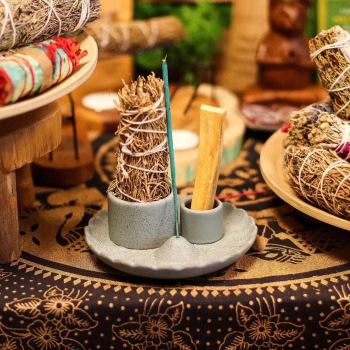 Gray ceramic smudge holder with two compartments holding sage bundles and a palo santo stick with lotus flower design