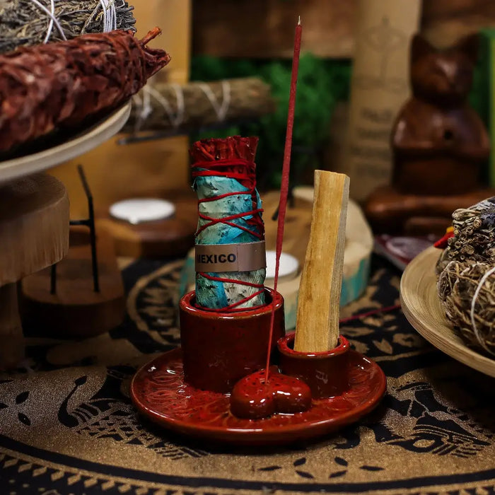 Red ceramic smudge holder with a mandala heart design holding a long palo santo stick and a wrapped sage bundle labeled mexico