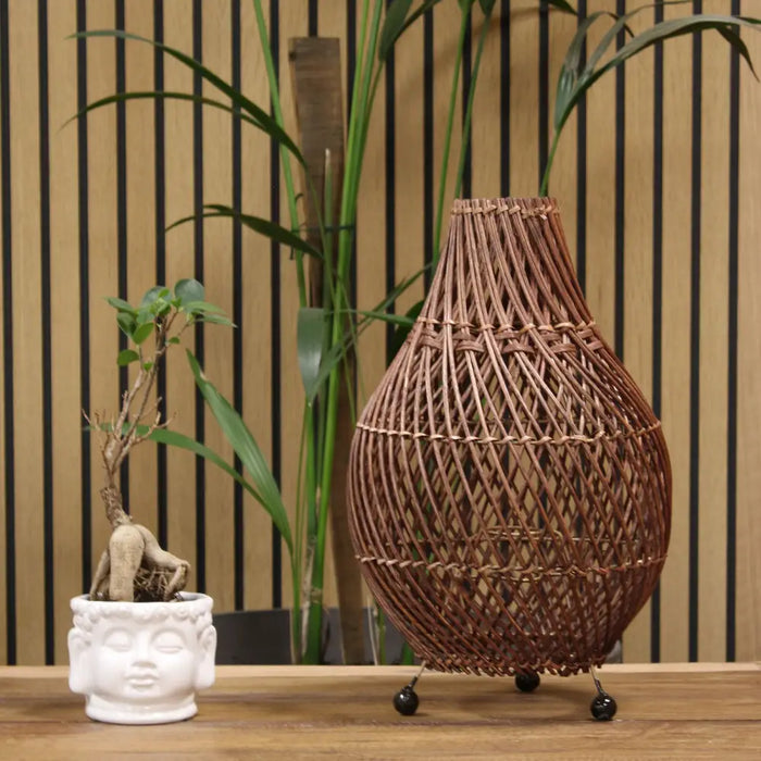 A dark brown rattan table lamp with a teardrop shape made of natural rattan and black spherical feet with plug fitting