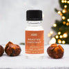 A clear glass bottle of roasted chestnut fragrance oil with a black cap and allergen declarations beside chestnuts on white surface