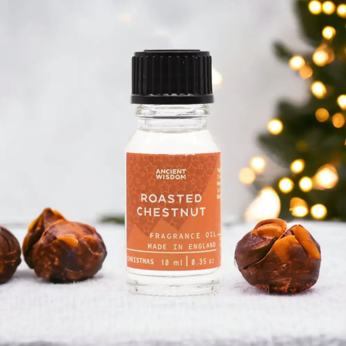 A clear glass bottle of roasted chestnut fragrance oil with a black cap and allergen declarations beside chestnuts on white surface