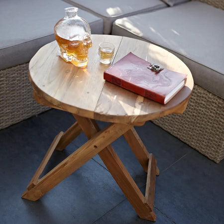 Clear glass skull-shaped decanter with amber liquid on round folding coffee table made of teak wood and recycled wood