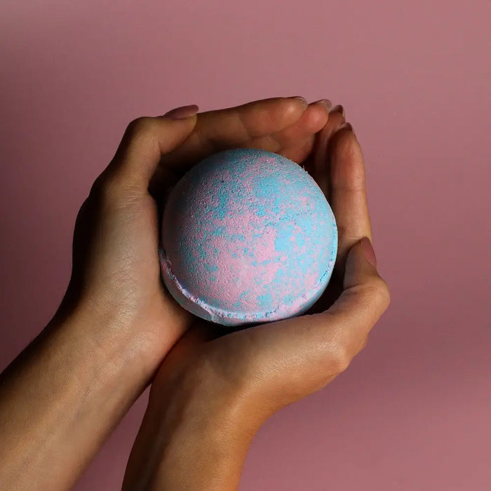 A round pastel blue and pink bath bomb with textured surface held in hands from a set of 3 baby powder bath bombs