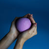 A soft purple spherical bath bomb held in two hands from a set of 3 bath bombs blackberry collection
