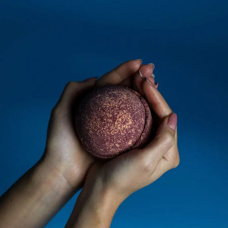 A maroon glitter bath bomb from a set of 3 bath bombs held gently in hands against a solid blue background