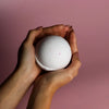 A white round coconut dream bath bomb with pink speckles held gently in two hands