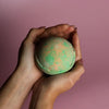 A round speckled green and orange bath bomb held in hands from the set of 3 bath bombs hawaiian mango