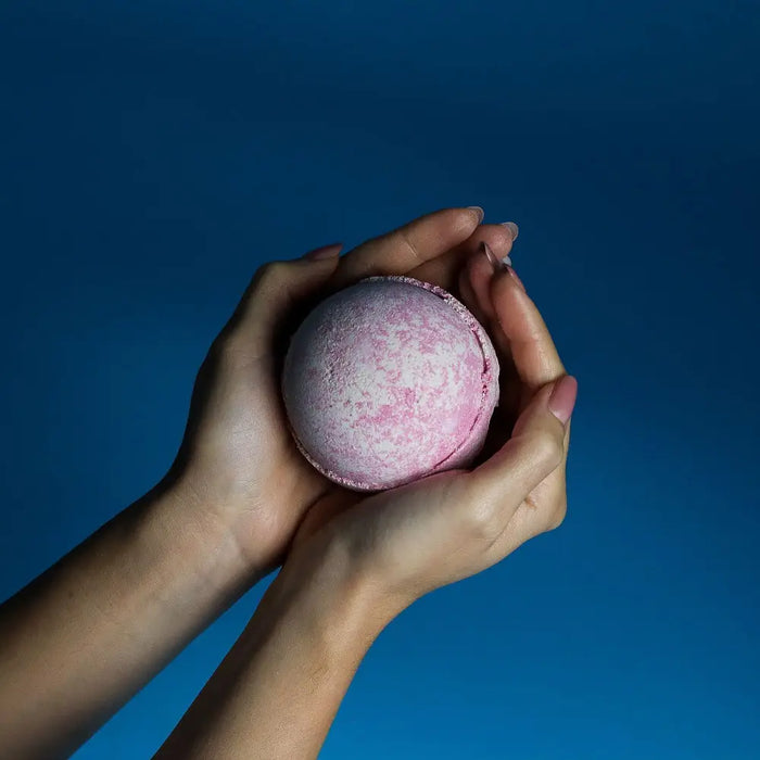 A soft pink round bath bomb with speckles held in hands against a blue background from the party girl bath bombs set