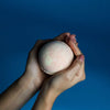 A soft pink peach sangria bath bomb with a textured surface held in two hands against a blue background