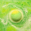 A green and yellow spherical bath bomb with tea tree scent floating in bubbly water from a set of 3 bath bombs