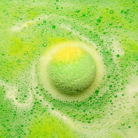 A green and yellow spherical bath bomb with tea tree scent floating in bubbly water from a set of 3 bath bombs