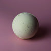 A pale green simply vanilla bath bomb with a rough surface and small dark specks in a set of 3 bath bombs