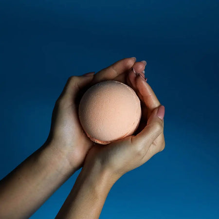 A soft peach-colored spherical bath bomb held in two hands from a set of 3 bath bombs tangerine and grapefruit