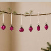 Small fuchsia mouth-blown glass baubles with metallic caps hang from white ribbons on a pine branch